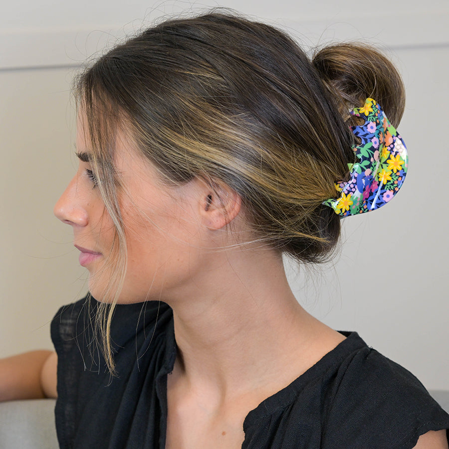 A woman with light brown hair in a bun, secured by a quality acrylic floral-patterned hair clip, sits on a chair, wearing a black top.