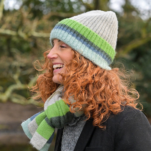 A woman with curly red hair, wearing a cosy knitted hat and coordinating striped scarf in green, blue, and grey, smiles outside.