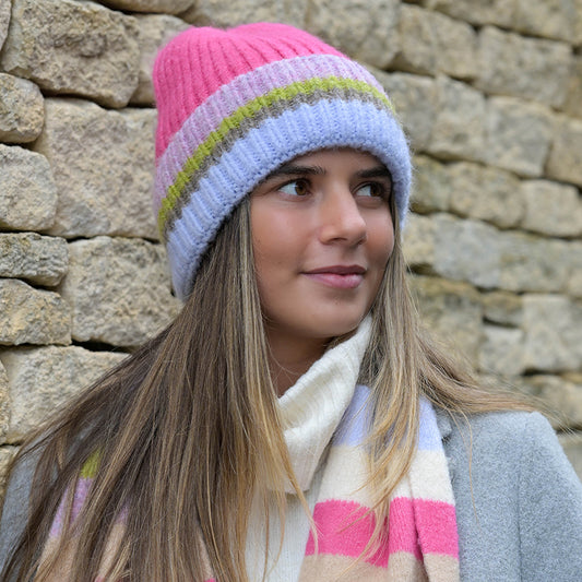 A young woman in a pink and mauve ribbed beanie hat and matching scarf stands before a stone wall, gazing to the side.