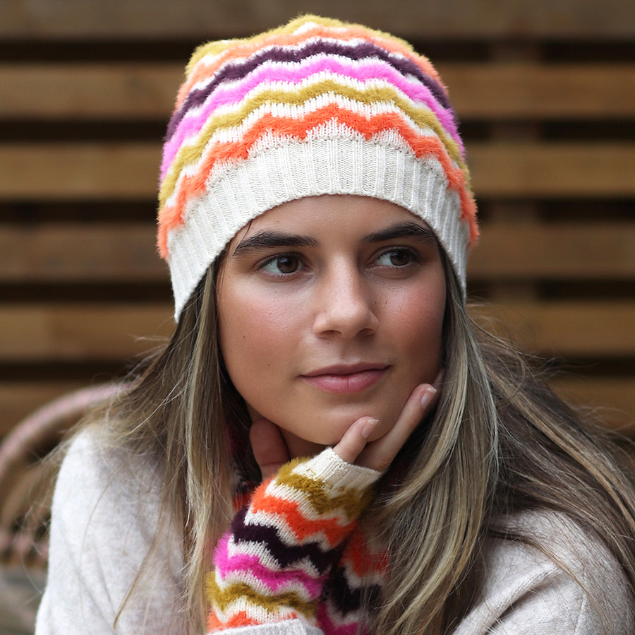 A woman wears a vegan-friendly chevron beanie hat and matching zigzag fingerless gloves, with a neutral backdrop of wooden planks.