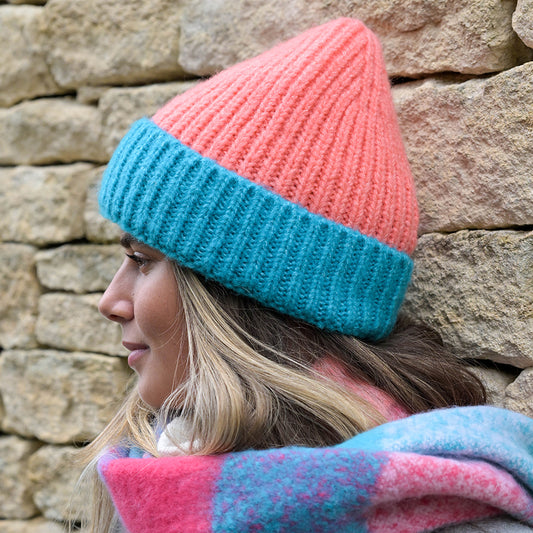 A woman in profile wears a cosy pink and blue hat and a colourful scarf, standing in front of a stone wall.