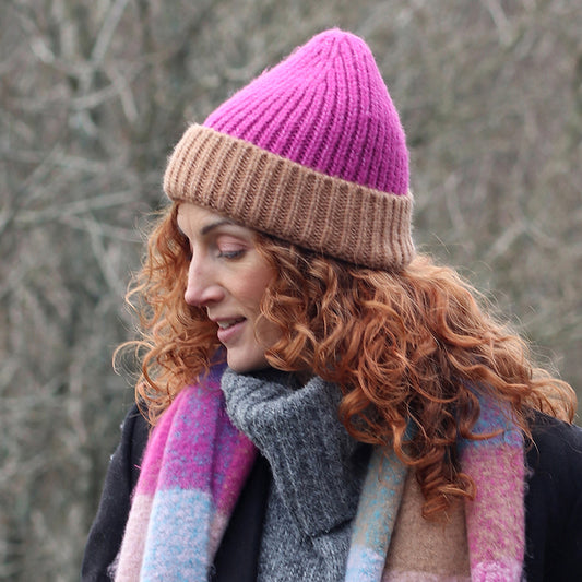 A woman with curly red hair wears a magenta and coffee beanie hat, a multicoloured scarf, and a grey coat as she stands outdoors in a wooded area.