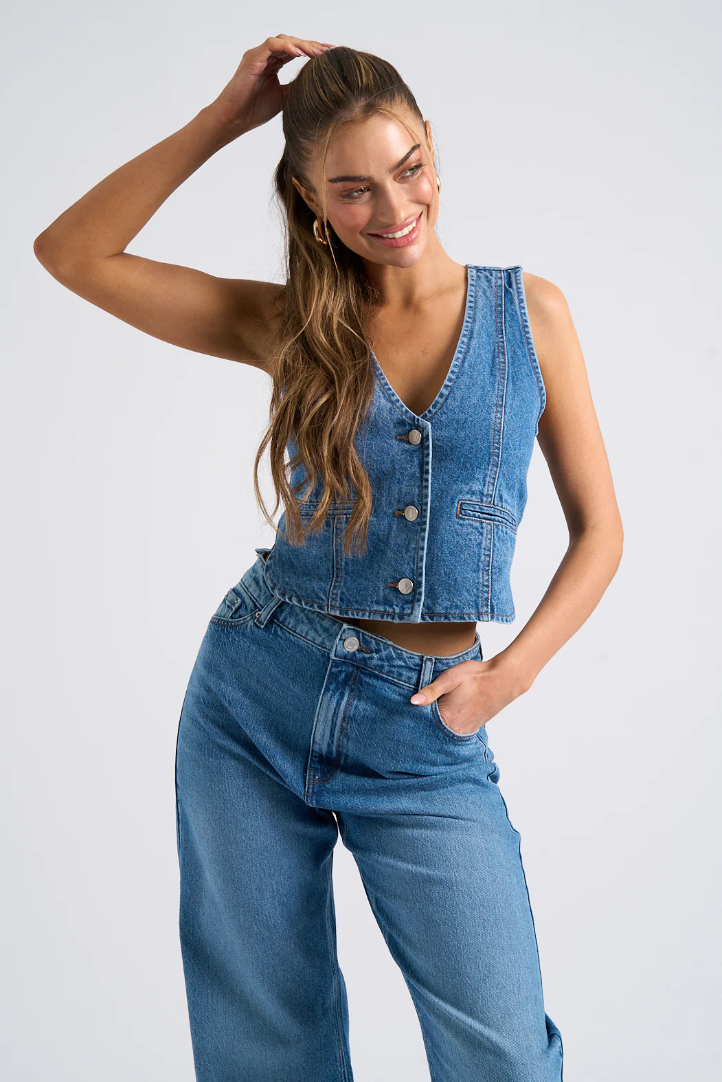 A woman with long hair, wearing a denim waistcoat and high-waisted jeans, is smiling. One hand rests on her hip while the other touches her hair against a plain background.
