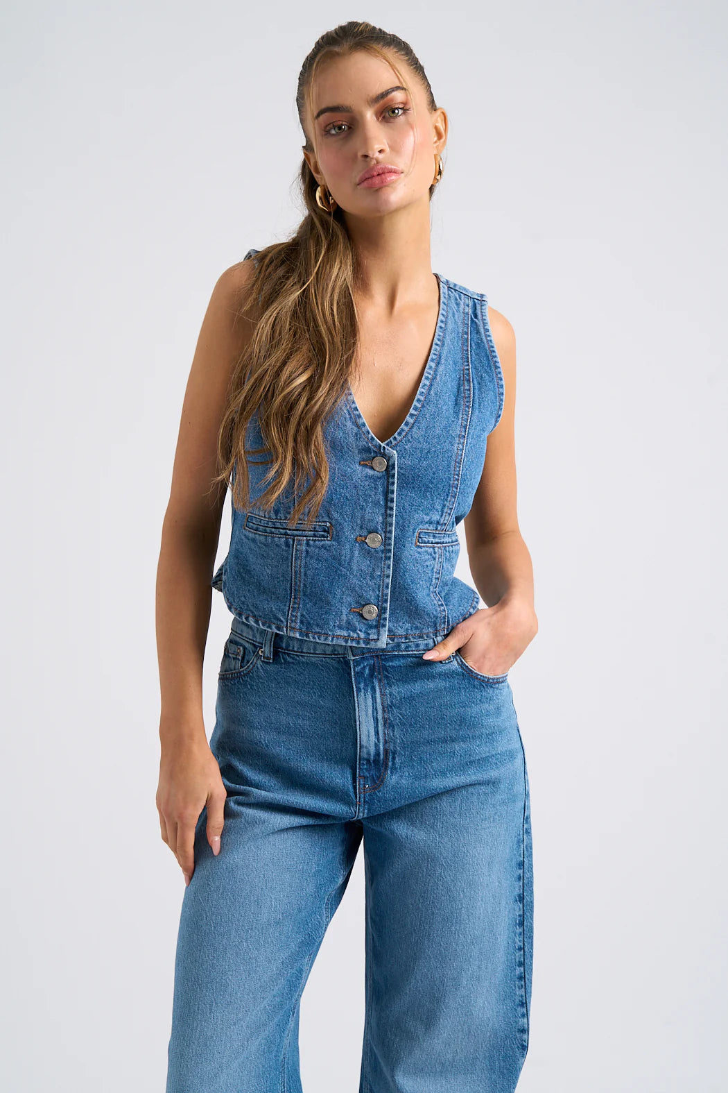 A person sports a denim waistcoat and high-waisted jeans, standing against a plain backdrop.