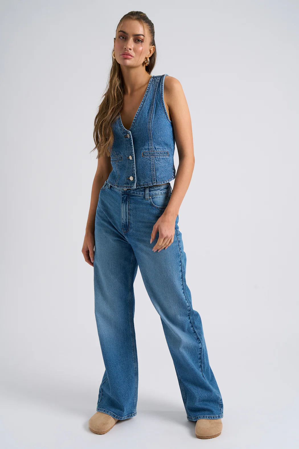 A person wearing a sleeveless denim waistcoat and high-waisted, wide-leg jeans with beige shoes stands against a plain background, showcasing the perfect denim staple look.