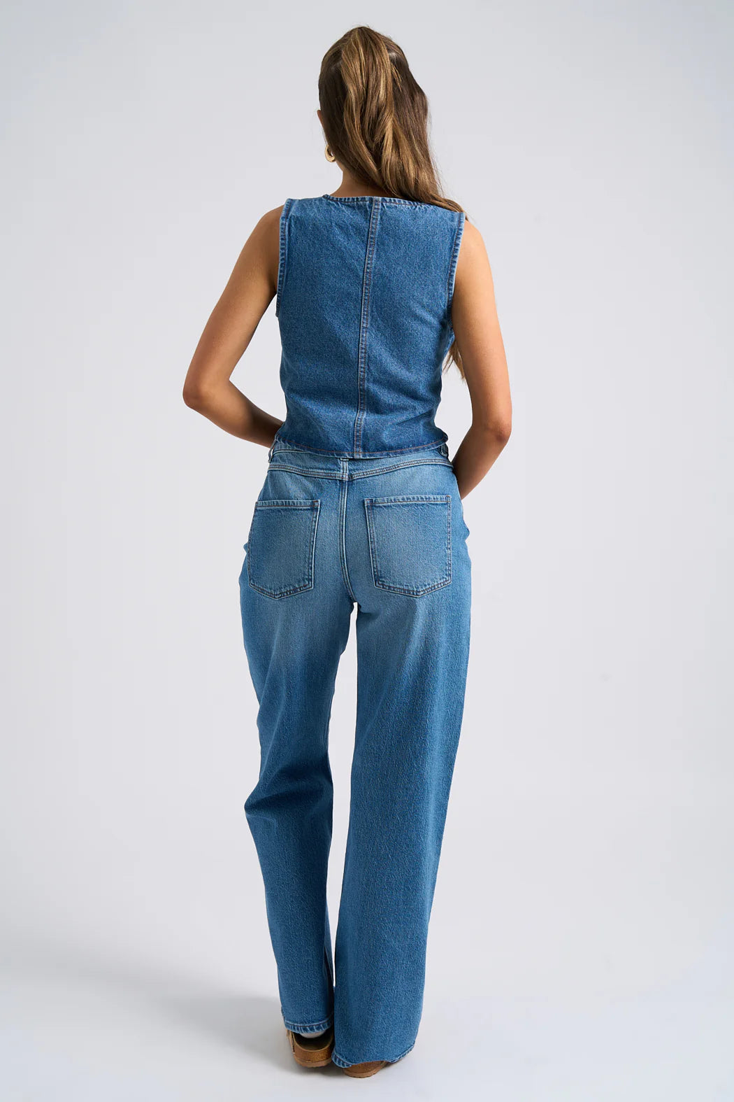 A person with long hair stands poised with their back turned, wearing a sleeveless denim jumpsuit that is truly a classic denim piece.