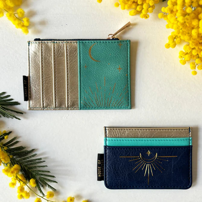 Two small, cruelty-free card holders with celestial designs—one turquoise and gold featuring a moon and stars, and one navy and gold with a sun—are set on a white surface adorned with yellow flowers.