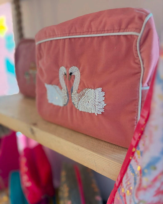 A Double Swan Large Pouch in pink velvet, featuring embroidered swans facing each other, is displayed on a wooden shelf, surrounded by other colourful fabric items and glass bead embellishments.