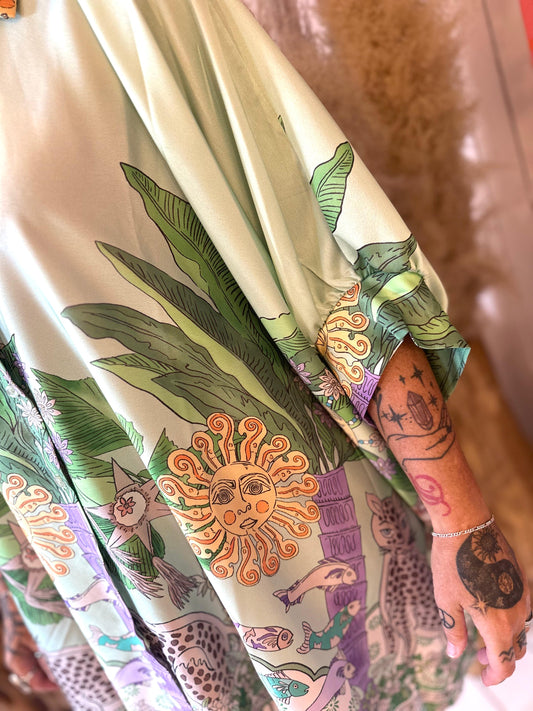 A person wearing a light green robe decorated with sun, plant, and animal patterns; their tattooed arm is partially visible.