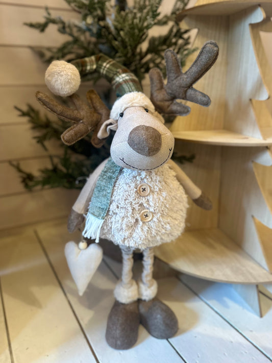 Plush reindeer figurine wearing a scarf and hat, holding a heart ornament, stands before a small artificial tree and wooden decorations—ideal festive décor for your home.