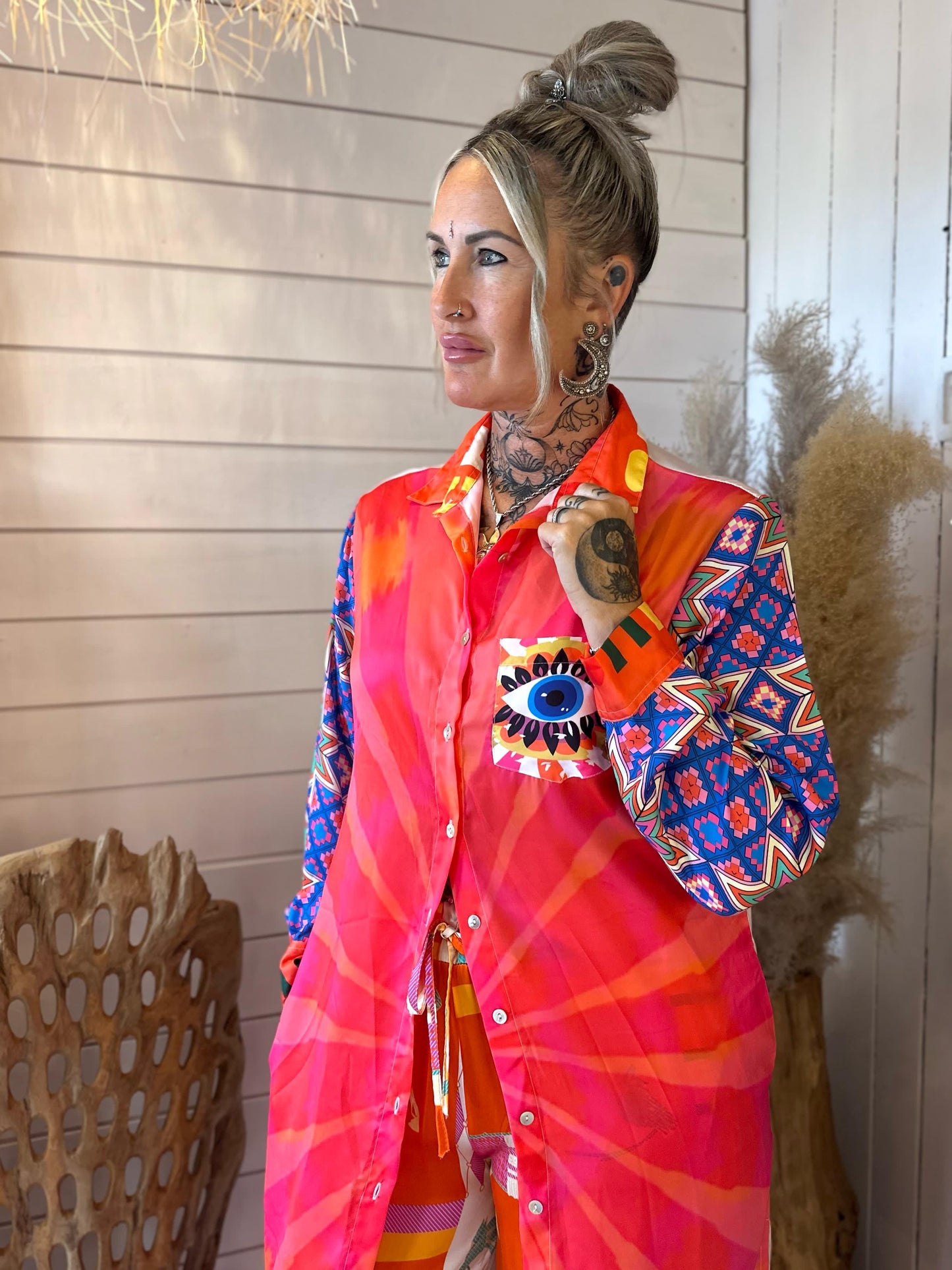 A woman with tattoos and an updo hairstyle wears a brightly patterned Eye Kaftan featuring a large eye design on the pocket, standing indoors near pampas grass and a wooden chair.