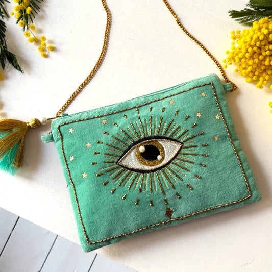 The Glimmer Crossbody Bag, a teal velvet handbag featuring a gold chain strap and embroidered eye, sits on a white surface adorned with yellow flowers—an elegant vegan-friendly accessory and an ideal Christmas gift.