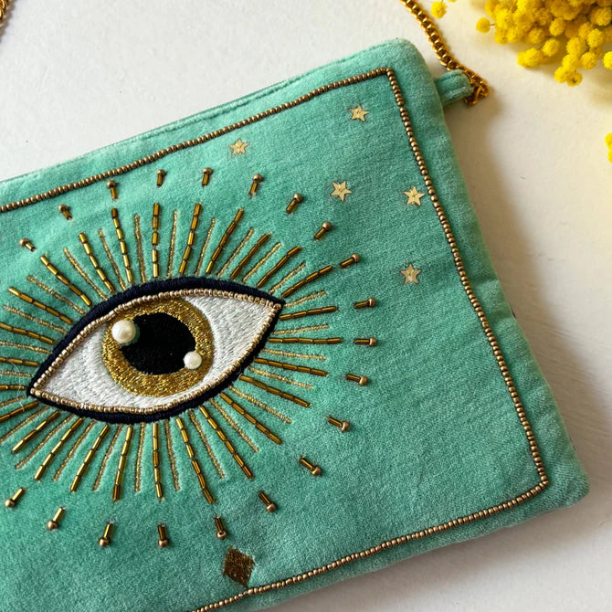 A teal fabric Glimmer crossbody bag with a gold chain, featuring an embroidered eye with gold rays and stars. Vegan-friendly accessory with yellow flowers in the top right—ideal as a distinctive Christmas present.