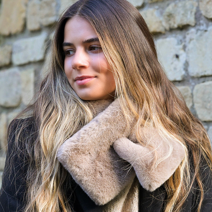 A woman with long, wavy hair wears a black coat and a taupe faux fur pull-through scarf, standing before a stone wall—a luxurious winter accessory.