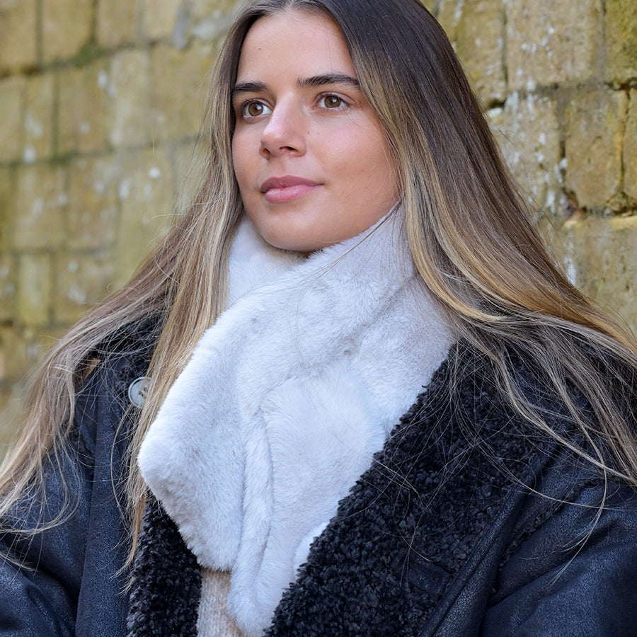 A woman with long hair, dressed in a black coat and a luxurious white pull-through scarf, stands in front of a stone wall.
