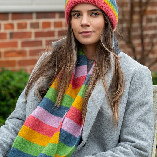 A woman in a grey coat, a mauve rainbow vegan scarf, and a matching knitted hat sits outdoors in front of a brick wall.