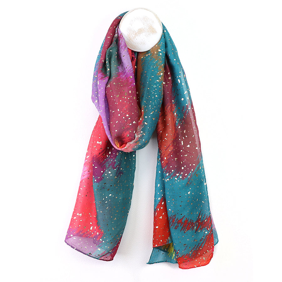 A colourful luxury scarf with a blend of teal, red, purple, and orange shades, featuring white speckles throughout and a subtle metallic foil overlay, hangs gracefully on a display stand.