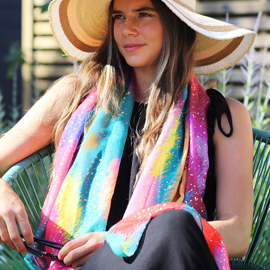Person sitting on a chair wearing a wide-brimmed hat, a luxury scarf with a metallic foil overlay, and holding sunglasses.