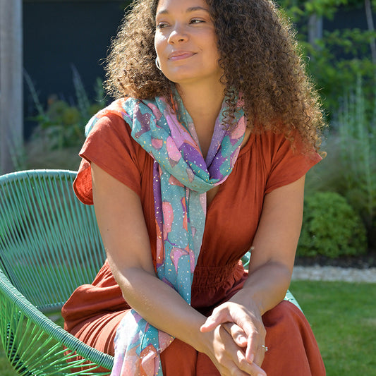 A person with curly hair is seated on a green chair outside, wearing a rust-coloured dress and an abstract butterfly-wing-patterned scarf, looking to the side.