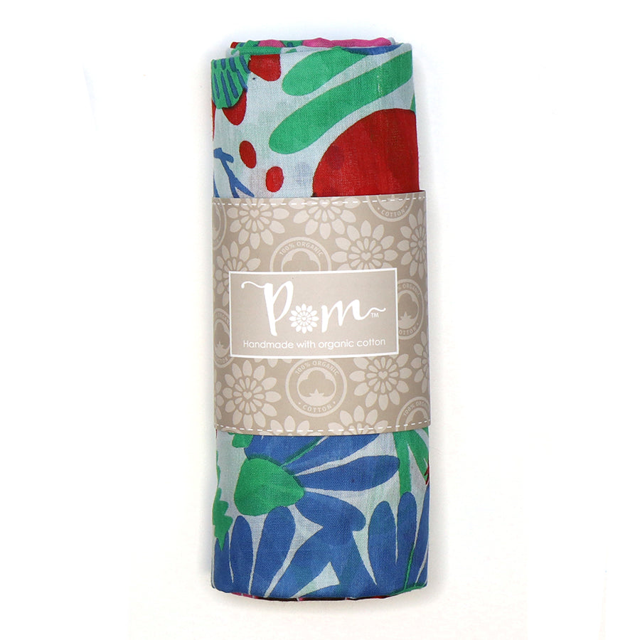 A rolled-up fabric with a vibrant meadow design in red, blue, and green is wrapped in a label reading Pom, Handmade with organic cotton.