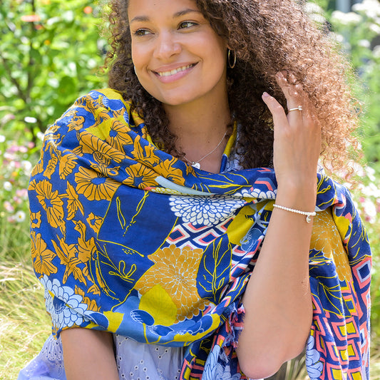 Woman smiling outdoors, wrapped in a vibrant Dahlia Multi Print shawl, basking in the suns glow amidst a lush garden background.