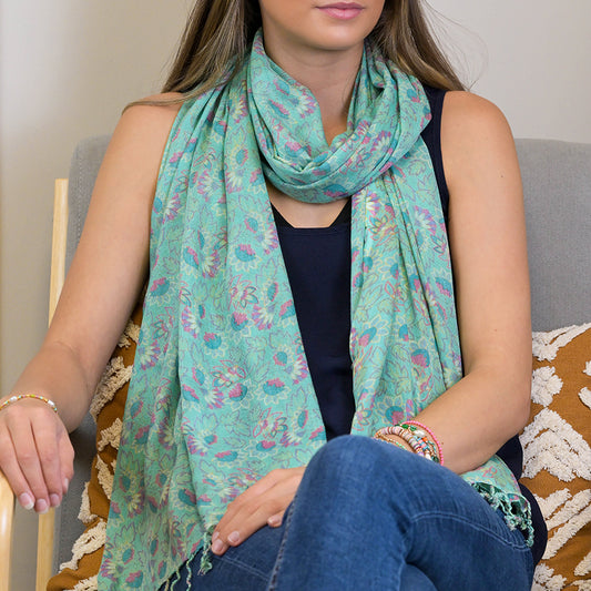 A woman is seated, wearing a luxurious floral scarf and a navy top, with her legs crossed and her hands resting on her lap.