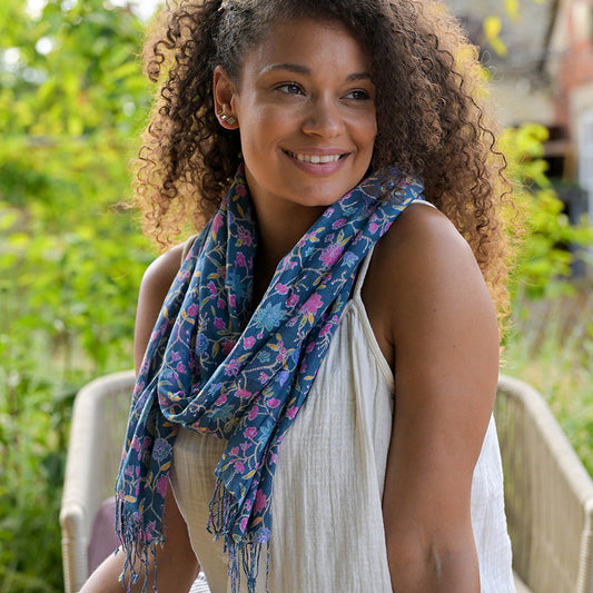 A woman seated outdoors, wearing a white top and a fashion-forward accessory—a designer scarf adorned with a beautiful blue floral pattern—smiling to the side with greenery in the background.