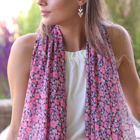 The woman, dressed in a white top and heart-shaped earrings, gazes to the side. A pink mix floral jasmine print scarf is elegantly draped around her neck, set against a backdrop of blurred greenery.