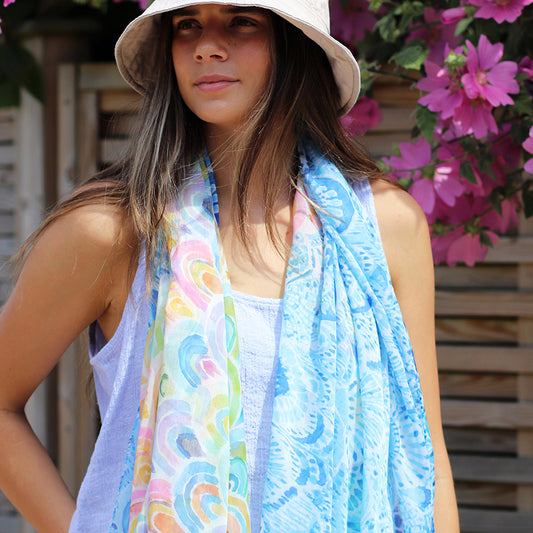 A woman in a light hat and blue sleeveless top stands near pink flowers, wearing a fashion-forward accessory—a colourful scarf with a watercolour rainbow print.
