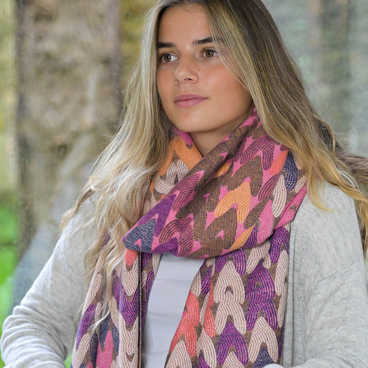 A woman with long hair wears a colourful, patterned reversible snug mix heart scarf and a light grey jumper. She stands indoors near a window, her environmental awareness reflected in the blurred outdoor background.