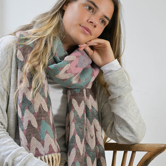 A woman with long hair, draped in a jacquard design scarf and wearing a light jumper, sits on a wooden chair with her hand resting on her cheek, looking pensively to the side.