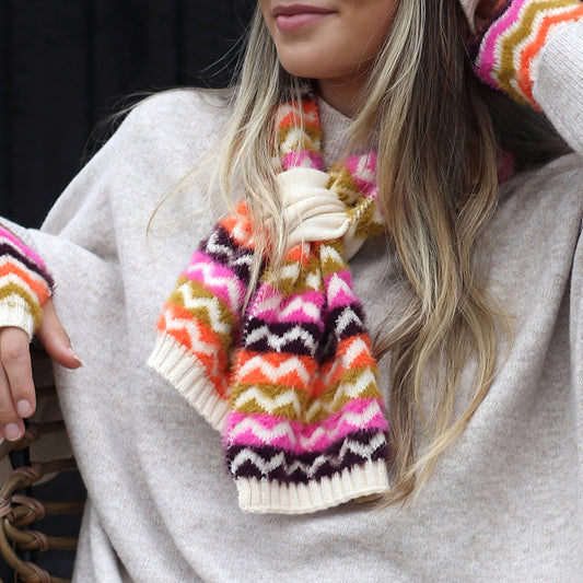 A woman in a beige jumper and a colourful chevron scarf with pink, orange, yellow, brown, and white patterns sits on a chair.