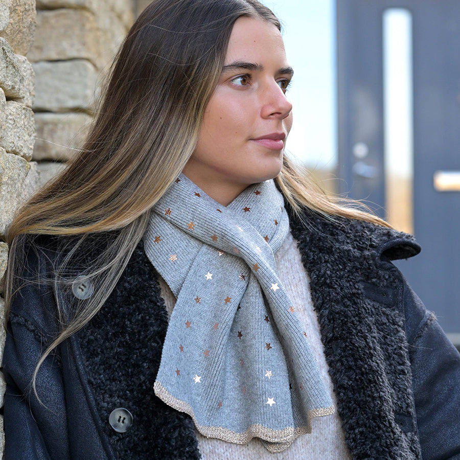 Woman with long hair wears a grey recycled-blend scarf featuring star patterns, a light jumper, and a black textured coat, standing beside a stone wall and gazing to the side.