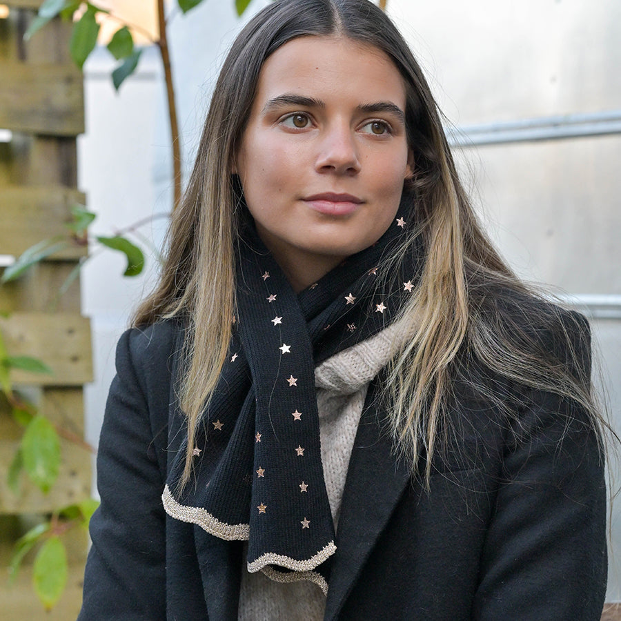 A woman with long brown hair, dressed in a black coat, beige jumper, and an eco-friendly scarf decorated with small gold stars, sits outdoors.