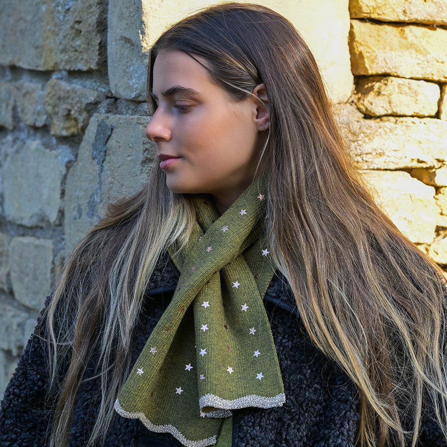 A young woman with long brown hair stands against a stone wall, dressed in a black coat and an olive green recycled blend scarf decorated with small white stars.