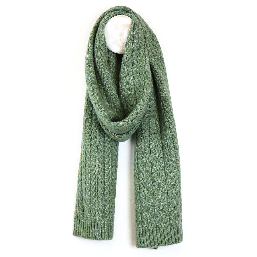A sage scarf, made from recycled vegan yarn, displays a chevron pattern and hangs over a white hook against a simple background.