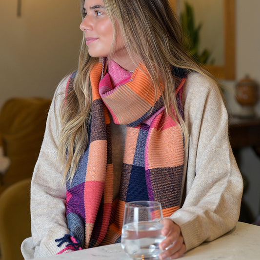 A woman with long hair is wearing a beige jumper and a rust and pink scarf, holding a glass of water and looking to the side indoors.