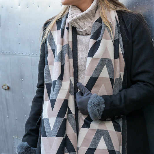 A person wearing a stylish chevron scarf, light polo-neck jumper, black coat, and gloves stands in front of a metal wall.