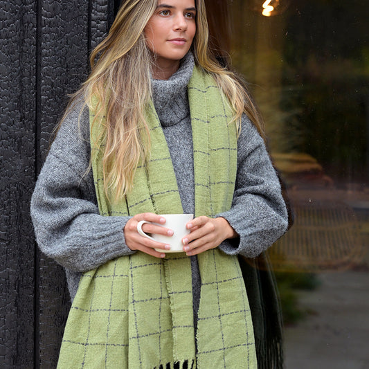 Woman in a grey jumper and cosy scarf stands by a dark wall, holding a white mug with both hands, gazing away from the camera.