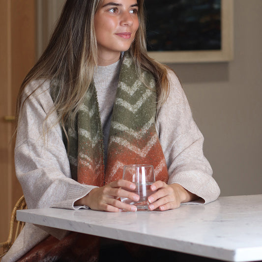 A woman wearing a cosy scarf and light jumper sits at a table, holding a clear glass of water with both hands.