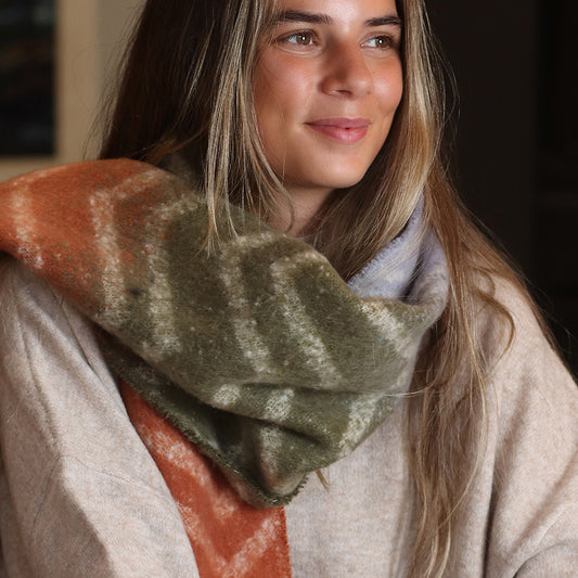 A woman with long brown hair wears a beige jumper and a large chevron scarf featuring green, orange, and white patterns. She is looking slightly to the side and smiling.