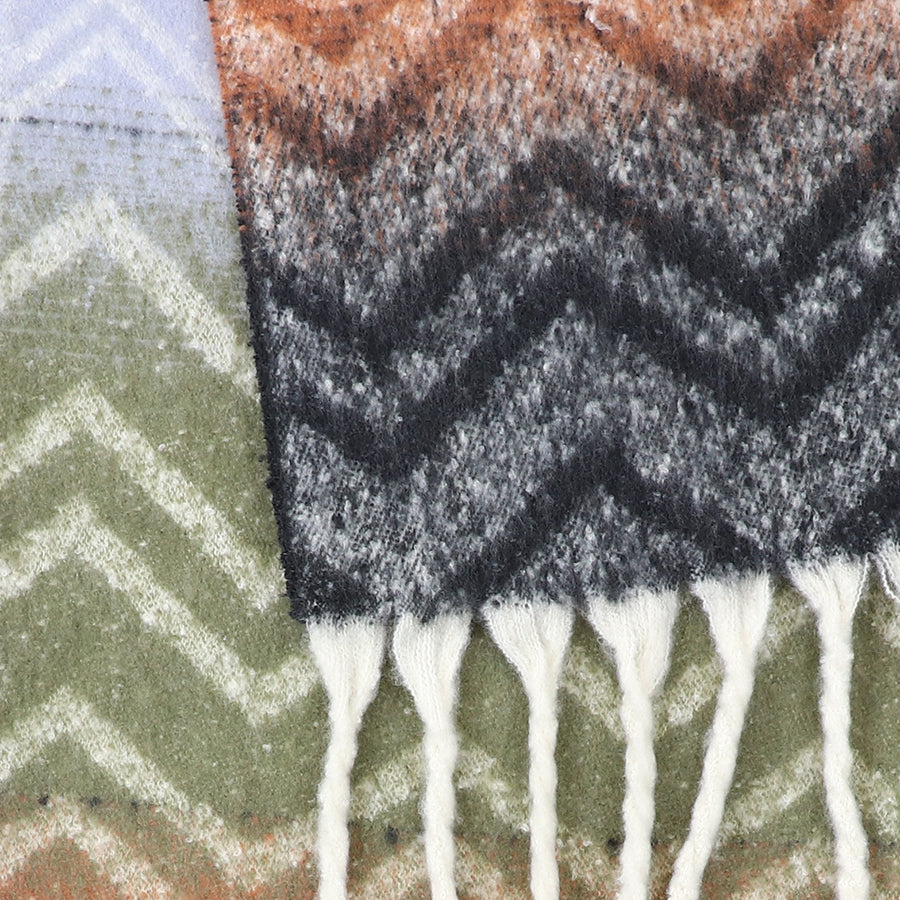 Close-up of a Chevron Scarf displaying a woven chevron pattern in green, brown, and black tones, with white fringed tassels along one edge.
