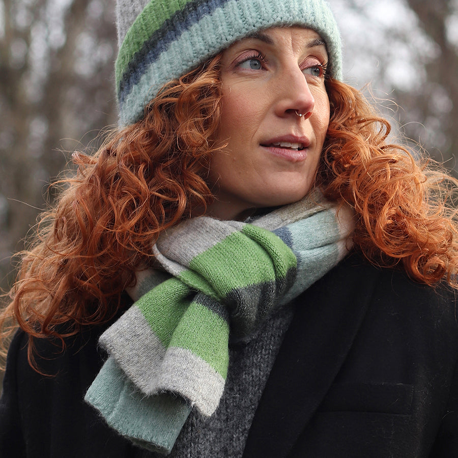 A person with curly red hair wearing a ribbed beanie and a green, grey, and white colourful scarf looks to the side outdoors in cold weather.