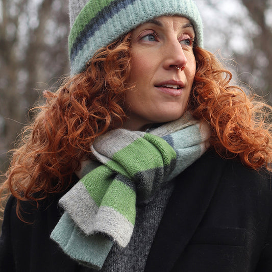 A person with curly red hair wearing a ribbed beanie and a green, grey, and white colourful scarf looks to the side outdoors in cold weather.