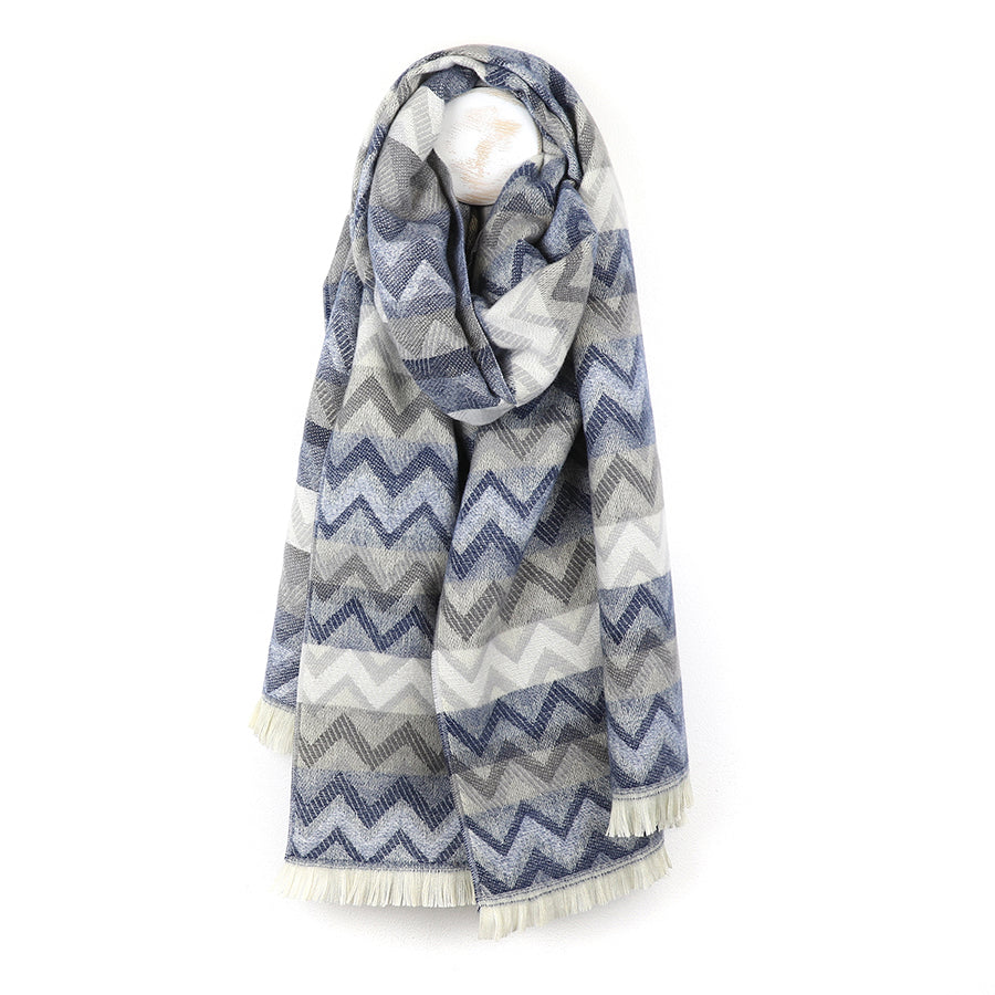 A long chevron scarf featuring a blue, grey, and white zigzag pattern is draped on a white background; the ends are finished with short fringe, making it a vibrant fashion accessory.