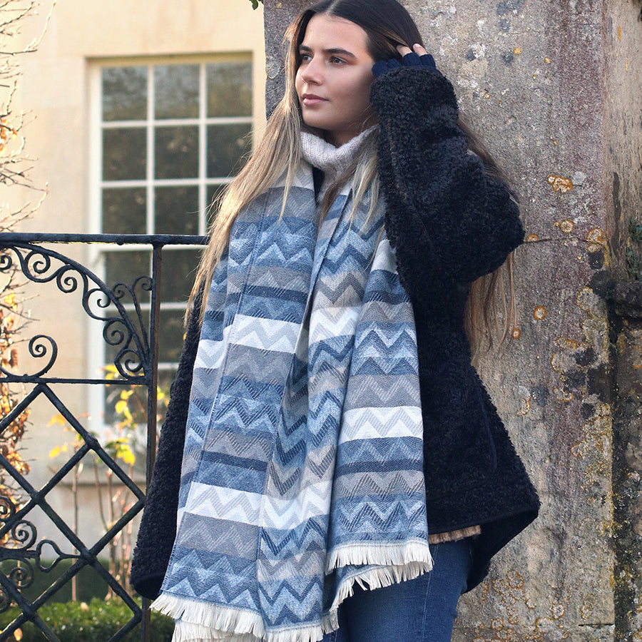 A woman with long hair, dressed in a thick black coat and a vibrant chevron scarf, stands outdoors near a stone wall and iron gate.
