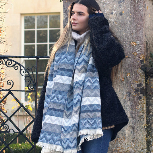 A woman with long hair, dressed in a thick black coat and a vibrant chevron scarf, stands outdoors near a stone wall and iron gate.