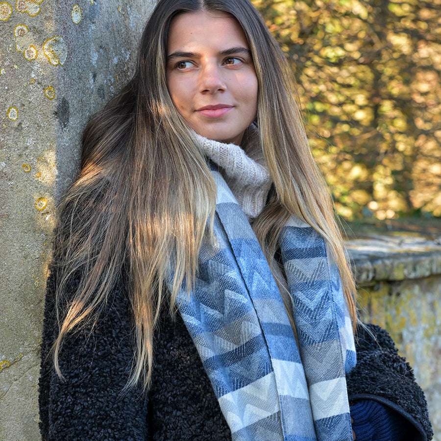 A woman with long brown hair, dressed in a knitted jumper, colourful scarf, and black coat, stands outdoors beside a stone wall with trees in the background.