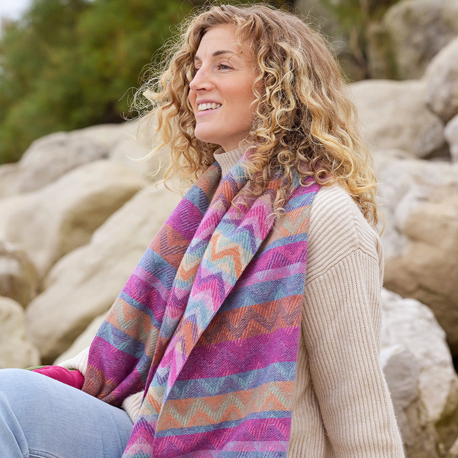 A woman with curly hair, dressed in a beige jumper and a vibrant fashion accessory—a colourful chevron scarf—sits on rocks outdoors, looking to the side and smiling.