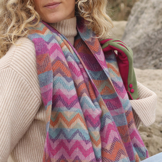 Woman in a beige polo-neck jumper, striking chevron scarf, and green gloves with pink edging, standing outdoors by rocks.